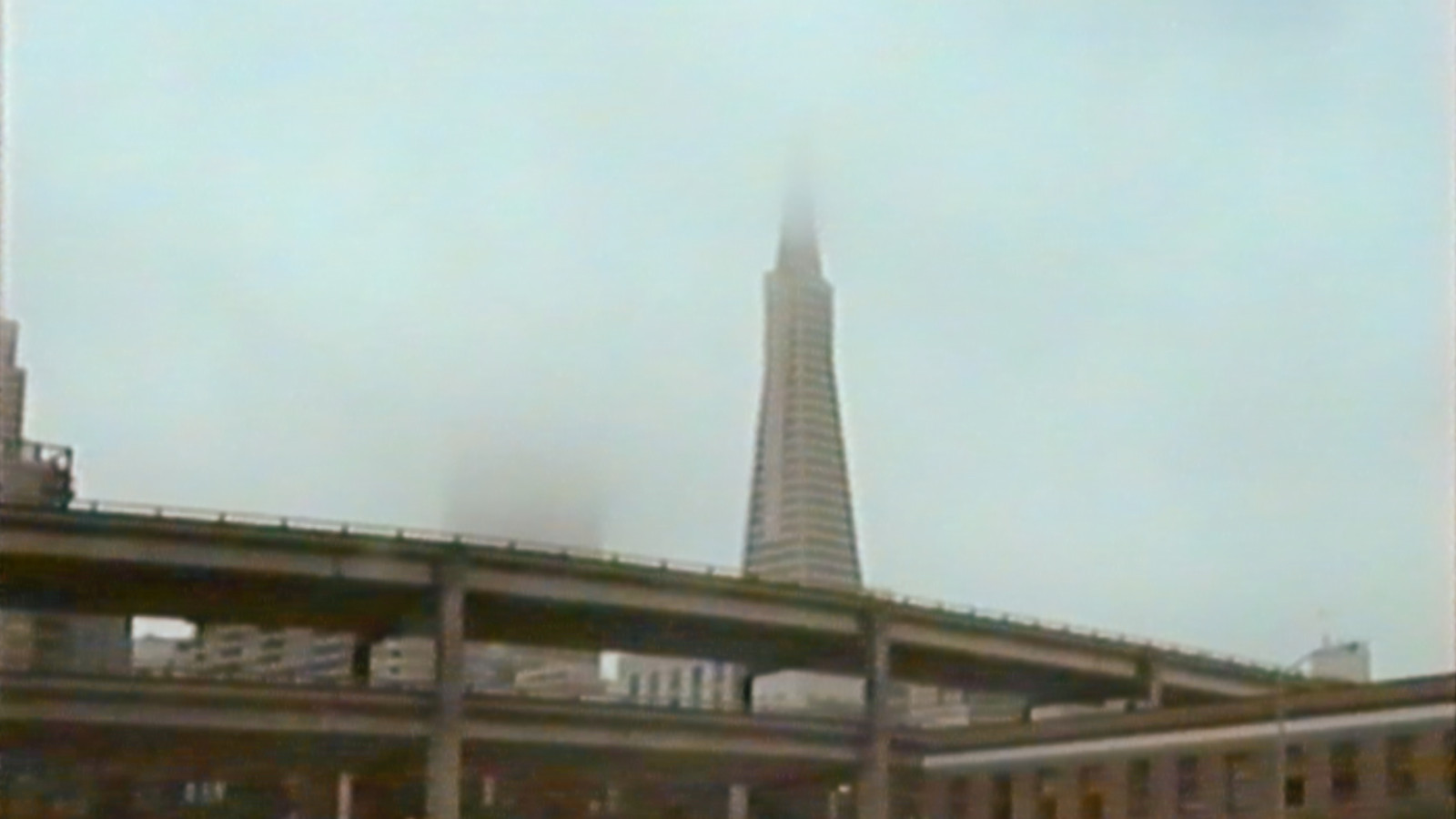 Still from a 1989 VHS home video, with the San Francisco Embarcadero Freeway in the foreground and the Transamerica Pyramid in the background, its tip hiding in the fog
