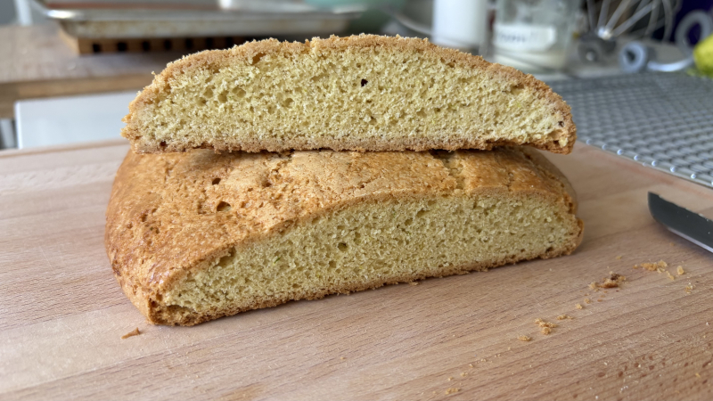 A flat loaf of baked brazadela (pâte sucrée cake) cut in half, one half on top of the other, with the cut end facing the viewer. The crust is crisp and lightly glazed with milk and sugar. The crumb is tight, with very small air bubbles