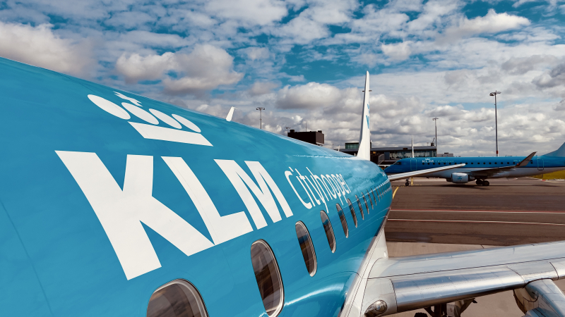 The fuselage of a KLM Cityhopper plane, with another KLM plane taxiing in the background against a KLM-blue sky dotted with small clouds